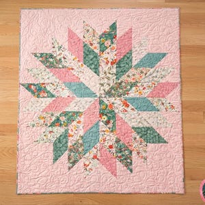May include: A handmade quilt featuring a starburst design. The quilt showcases a central star pattern with alternating segments of pink, teal, and floral fabrics. The background is a solid pink, and the quilt has a quilted texture.