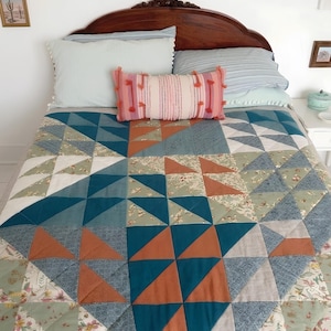 Puede incluir: Una cama con un edredón de retazos con triángulos turquesa, naranja y blancos. El edredón está rematado con almohadas azul claro y a rayas, y una almohada decorativa rosa con borlas. Un cabecero de madera oscura completa la escena.