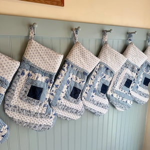 以下が含まれることがあります： さまざまな青と白の模様（花柄やギンガムチェックなど）の手作りのクリスマスストッキングの列。各ストッキングはキルトのような外観で、水色の壁に掛けられています。お祝いの休日の装飾です。