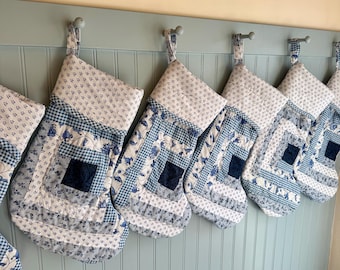 Könnte beinhalten: Eine Reihe handgefertigter Weihnachtsstrümpfe in verschiedenen blau-weißen Mustern, darunter Blumen- und Vichy-Designs. Jeder Strumpf hat ein gestepptes Aussehen und hängt an einer hellblauen Wand. Eine festliche Weihnachtsdekoration.