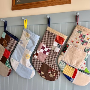 Könnte beinhalten: Vier Patchwork-Weihnachtsstrümpfe hängen an einer hellblauen Wand. Jeder Strumpf hat ein anderes Muster, darunter florale, karierte und einfarbige Stoffquadrate. Die Strümpfe sind an Schlaufen aus blauem, gelbem und rotem Band aufgehängt.