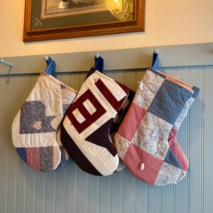 以下が含まれることがあります： 青いペグに吊るされた、3つのキルトのクリスマスストッキング。ストッキングは、赤、青、白など、さまざまな色のパッチワークデザインが特徴です。1つのストッキングには、文字「B」がアップリケされています。