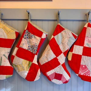 Puede incluir: Cuatro calcetines navideños hechos de tela de patchwork acolchada con una variedad de patrones florales y geométricos en rojo, blanco, amarillo y azul. Cada calcetín tiene un lazo para colgar y costuras visibles en toda la superficie.