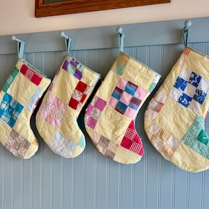 Puede incluir: Cuatro calcetines de Navidad hechos a mano colgados en una pared azul claro. Cada calcetín tiene un fondo amarillo con cuadrados de patchwork coloridos. Los calcetines están suspendidos por cintas azul claro de pequeños ganchos redondos.