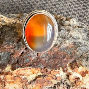 Peut inclure: Une bague en argent ornée d'une grande pierre cabochon de forme ovale. La pierre présente une surface lisse et polie avec un dégradé de tons orange et gris translucides, sertie dans une monture simple en métal argenté poli.