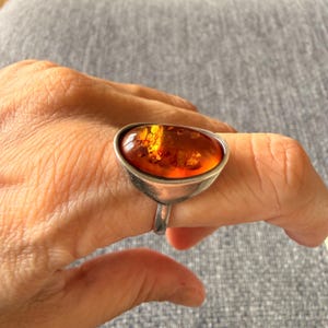 Könnte beinhalten: Ein Ring mit einem großen, ovalen, durchscheinenden Bernstein mit goldenen Einschlüssen. Der Stein ist in eine glatte, polierte, silberfarbene Metallfassung eingefasst, die in eine einfache, abgerundete Ringschiene übergeht.
