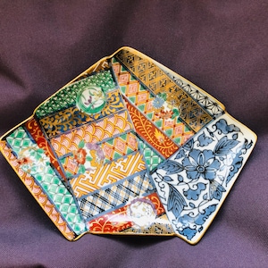 May include: A decorative, multi-patterned ceramic plate with a gold trim. The plate features a variety of colourful designs, including floral, geometric, and abstract patterns in shades of blue, green, orange, and yellow. The plate has a unique, angular shape.