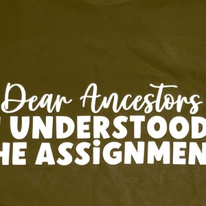 Könnte beinhalten: Olivgrünes T-Shirt mit dem weißen Text "Dear Ancestors I UNDERSTOOD THE ASSIGNMENT" in einer fetten, verspielten Schriftart. Das Design des Shirts ist einfach und direkt.