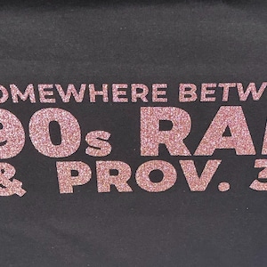 Puede incluir: Camiseta negra con el texto "SOMEWHERE BETWEEN 90s RAP & PROV. 31" en letras rosas brillantes. El texto está centrado y ocupa la mayor parte de la superficie.