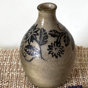 May include: A handcrafted, beige ceramic vase with a dark floral design. The vase has a rounded body and a narrow neck. The floral pattern includes stylized flowers and leaves. The vase is displayed on a woven surface.