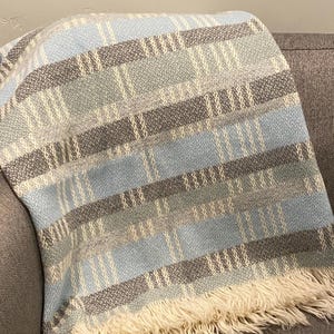 Puede incluir: Una manta tejida con un patrón de rayas azules, grises y crema. La manta tiene un borde con flecos y está colocada sobre un sofá gris. La paleta de colores es neutra y la textura parece suave.