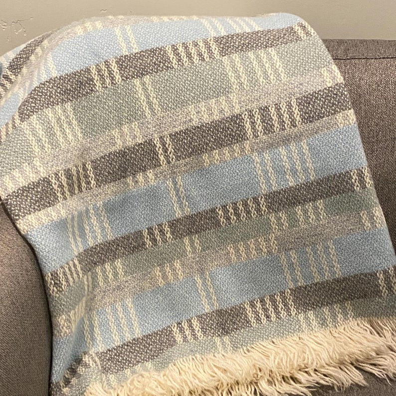 Op de afbeelding: Een geweven sprei met een blauw, grijs en cr&egrave;mekleurig streepjespatroon. De sprei heeft een gefranjerde rand en is over een grijze bank gedrapeerd. Het kleurenpalet is neutraal en de textuur lijkt zacht.