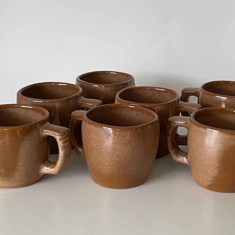 Pode incluir: Uma cole&ccedil;&atilde;o de seis canecas de cer&acirc;mica com um esmalte castanho brilhante e salpicado. Cada caneca apresenta um corpo arredondado e uma pega espessa e robusta fixada na lateral. As canecas est&atilde;o dispostas em grupo, exibindo a sua cor terrosa uniforme e um acabamento suave e refletor em todas as superf&iacute;cies.