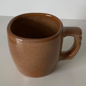 Pode incluir: Uma caneca de cer&acirc;mica castanha com um acabamento liso e brilhante e uma textura salpicada. A caneca apresenta um corpo arredondado e uma pega grossa e robusta com tr&ecirc;s cristas horizontais distintas na extremidade exterior.