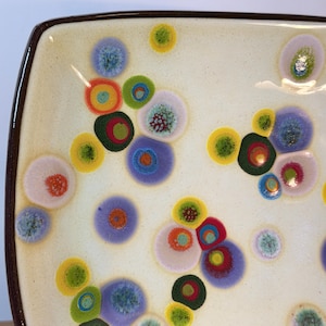 May include: A square ceramic plate with a cream-colored base featuring a scattered pattern of multi-colored, circular, and organic-shaped spots in shades of purple, blue, green, yellow, orange, and pink, finished with a dark brown rim.