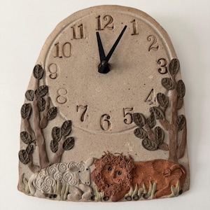 May include: A beige ceramic wall clock with a rounded top, featuring embossed numbers from 1 to 12 and black hands. The base displays a sculpted scene of a white sheep and a brown lion resting among trees with textured leaves on a speckled surface.