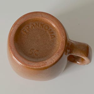 Pode incluir: Uma caneca de cer&acirc;mica castanha vista de baixo, com um acabamento brilhante e uma pega lateral. A base est&aacute; gravada com o texto FRANKOMA e os caracteres 5C, apresentando uma &aacute;rea circular rebaixada e uma forma lisa e arredondada.