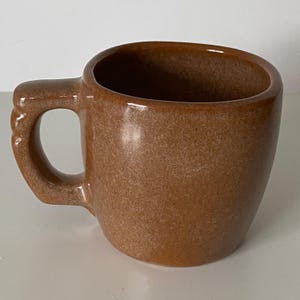 Pode incluir: Uma caneca de cer&acirc;mica castanha s&oacute;lida com um acabamento liso e salpicado, com uma pega grossa e resistente. A caneca apresenta um corpo cil&iacute;ndrico arredondado com uma abertura larga e um tom terroso consistente em toda a superf&iacute;cie.