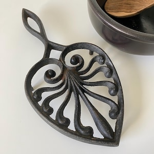 May include: A black cast iron trivet with a leaf-like design and a handle. The trivet features an intricate, openwork pattern of swirls and curves. A dark bowl and wooden spoon are in the background.
