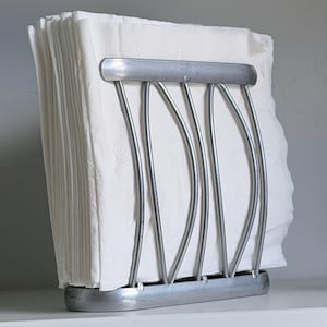 May include: A silver-toned metal napkin holder with a curved, vertical bar design. The holder features a solid base and a top horizontal bar, securely containing a stack of white paper napkins with a subtle embossed pattern on their surface.