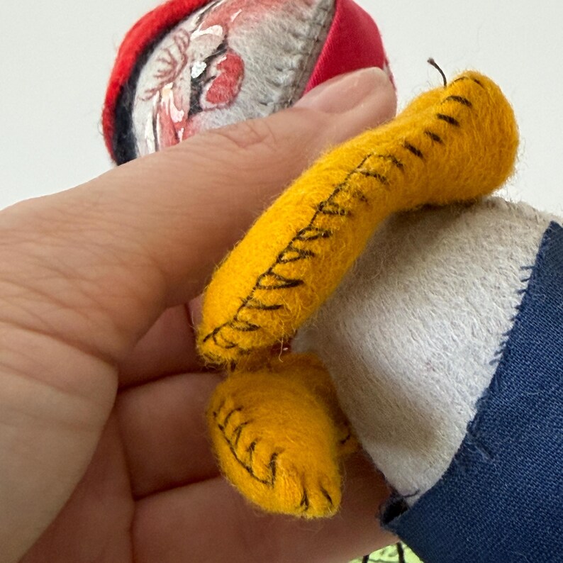 K&ouml;nnte beinhalten: Nahaufnahme einer handgefertigten Filzpuppe mit roter M&uuml;tze und gelbem Filzarm und -fu&szlig;. Arm und Fu&szlig; der Puppe sind mit schwarzem Faden gen&auml;ht. Der Kopf der Puppe hat ein gedrucktes Design mit roten und wei&szlig;en Farben.