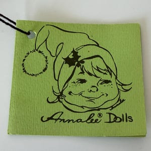 K&ouml;nnte beinhalten: Ein gr&uuml;nes Etikett mit schwarzem Faden und einer Cartoon-Illustration eines l&auml;chelnden Kindes mit Weihnachtsm&uuml;tze. Auf dem Etikett steht "Annalee Dolls" in Schreibschrift.