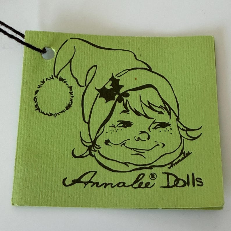 K&ouml;nnte beinhalten: Ein gr&uuml;nes Etikett mit schwarzem Faden und einer Cartoon-Illustration eines l&auml;chelnden Kindes mit Weihnachtsm&uuml;tze. Auf dem Etikett steht "Annalee Dolls" in Schreibschrift.