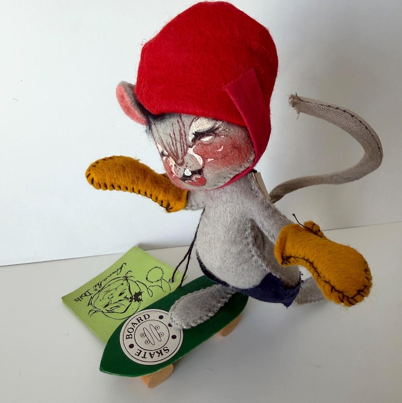K&ouml;nnte beinhalten: Eine handgefertigte Vintage-Mauspuppe mit roter M&uuml;tze, gelben Handschuhen und blauen Shorts, die auf einem gr&uuml;nen Brett skatet. Die Puppe hat ein bemaltes Gesicht und h&auml;lt ein kleines gr&uuml;nes Etikett mit Text und einer Cartoon-Maus-Illustration.