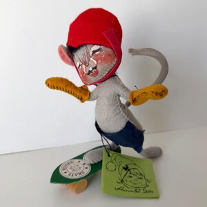 K&ouml;nnte beinhalten: Handgefertigte Mauspuppe mit roter M&uuml;tze, gelben Handschuhen und blauen Shorts, die auf einem gr&uuml;nen Board skateboardet. Die Puppe hat ein bemaltes Gesicht und ein Etikett mit der Aufschrift "Annale Dolls".