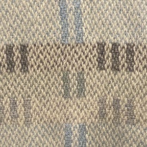 Op de afbeelding: Close-up van een geweven stof met een getextureerde uitstraling. De stof heeft een neutraal kleurenpalet met een lichtbeige basis en accenten van grijs en lichtblauw in een ruitpatroon. De weving is strak en gedetailleerd.
