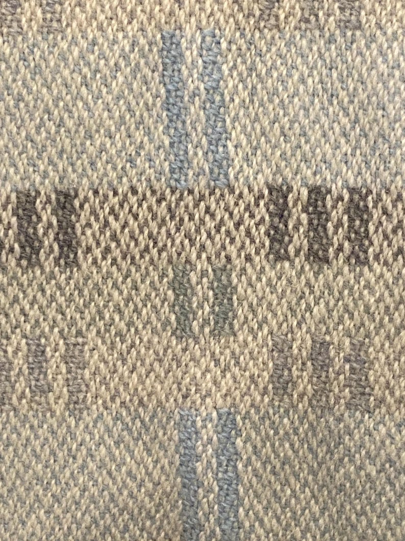 Op de afbeelding: Close-up van een geweven stof met een getextureerde uitstraling. De stof heeft een neutraal kleurenpalet met een lichtbeige basis en accenten van grijs en lichtblauw in een ruitpatroon. De weving is strak en gedetailleerd.