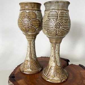 May include: Two handcrafted ceramic goblets with a floral design. The goblets are beige and brown, with a tall stem and a wide base. The floral design is embossed on the goblet's body. The goblets are approximately 8 inches tall.