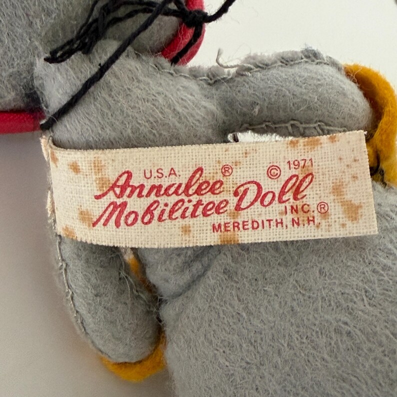 K&ouml;nnte beinhalten: Nahaufnahme einer Vintage-Annalee-Puppe. Die Puppe ist grau mit schwarzen Haaren und einem roten und gelben Akzent. Auf dem Etikett steht "Annalee Doll Mobilitee Inc. Meredith, N.H. U.S.A. &copy; 1971".
