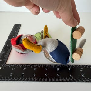 K&ouml;nnte beinhalten: Ein handgefertigtes Stofftier mit rotem Hut, gelbem Schnabel und blau-grauem K&ouml;rper. Das Spielzeug hat ein gr&uuml;nes Skateboard mit Holzr&auml;dern. Ein Lineal liegt neben dem Spielzeug und misst in Zentimetern.
