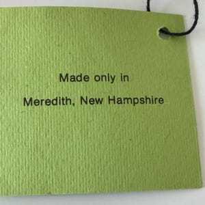 K&ouml;nnte beinhalten: Ein gr&uuml;nes rechteckiges Etikett mit dem Text "Made only in Meredith, New Hampshire" in Schwarz gedruckt. Das Etikett hat eine strukturierte Oberfl&auml;che und eine schwarze Schnur oben.