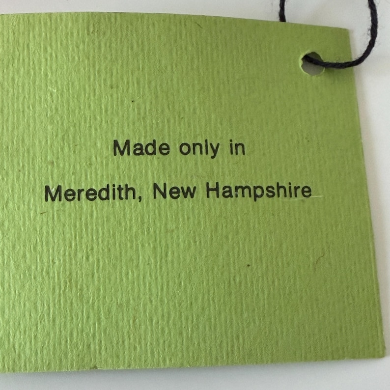 K&ouml;nnte beinhalten: Ein gr&uuml;nes rechteckiges Etikett mit dem Text "Made only in Meredith, New Hampshire" in Schwarz gedruckt. Das Etikett hat eine strukturierte Oberfl&auml;che und eine schwarze Schnur oben.