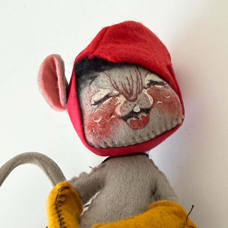 K&ouml;nnte beinhalten: Eine Vintage-Mauspuppe aus Filz mit roter Kapuze, grauem K&ouml;rper und gelben Akzenten. Die Maus hat ein l&auml;chelndes Gesicht mit rosigen Wangen und einem zahnigen Grinsen. Die Ohren der Puppe sind rosa, und sie scheint ein graues, gebogenes Objekt zu halten.