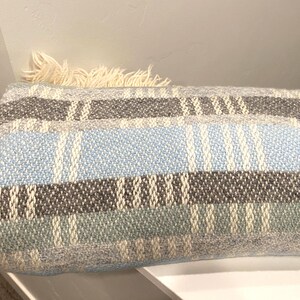 Op de afbeelding: Een rechthoekig geweven kussen met een gestreept patroon in tinten blauw, grijs en cr&egrave;me. Het kussen heeft een franje detail aan &eacute;&eacute;n uiteinde. Het kussen ligt op een wit oppervlak.