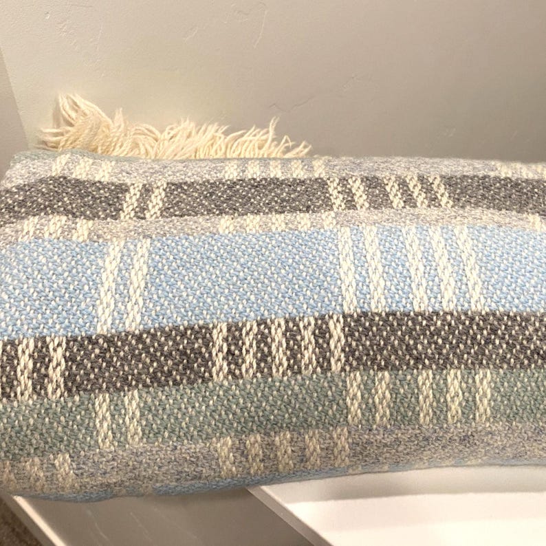 Op de afbeelding: Een rechthoekig geweven kussen met een gestreept patroon in tinten blauw, grijs en cr&egrave;me. Het kussen heeft een franje detail aan &eacute;&eacute;n uiteinde. Het kussen ligt op een wit oppervlak.