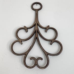 May include: A decorative, wrought iron wall hanging with a dark brown finish. The design features a central stem with a loop at the top for hanging, and stylized, symmetrical scrollwork. The piece is approximately 10 inches in height.