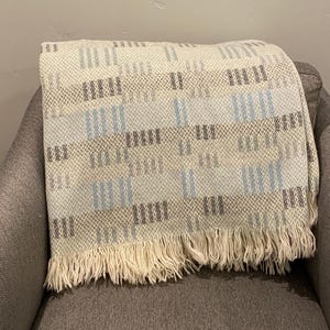 Op de afbeelding: Een geweven sprei met een cr&egrave;mekleurige basis en een geometrisch patroon in grijstinten en lichtblauw. De deken heeft een gefranjerde rand en is over een grijze fauteuil gedrapeerd.