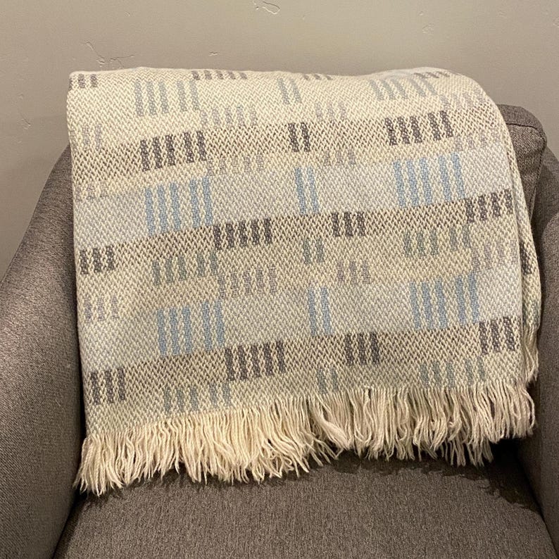 Op de afbeelding: Een geweven sprei met een cr&egrave;mekleurige basis en een geometrisch patroon in grijstinten en lichtblauw. De deken heeft een gefranjerde rand en is over een grijze fauteuil gedrapeerd.