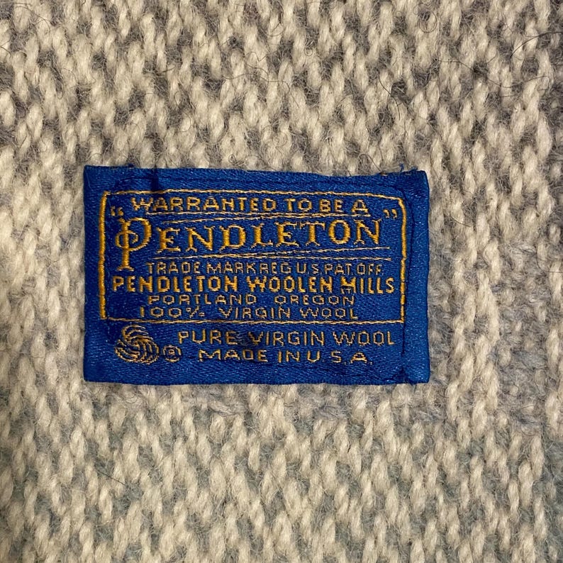 Op de afbeelding: Close-up van een blauw Pendleton-label met gouden letters, genaaid op een grijs en witte wollen stof. Het label luidt "Warranted to be a Pendleton" en bevat details over het merk en het materiaal.