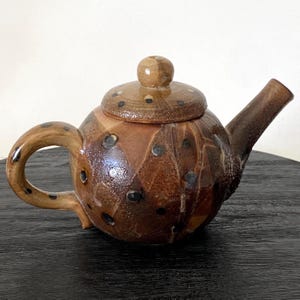 May include: A handcrafted ceramic teapot with a round body, a curved handle, and a spout. The teapot is primarily brown with darker brown and black polka dots. The lid has a small knob and also features polka dots.