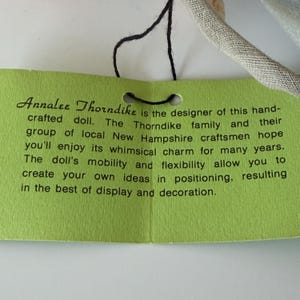 K&ouml;nnte beinhalten: Ein gr&uuml;nes Etikett mit schwarzem Text, auf dem steht: "Annalee Thorndike ist die Designerin dieser handgefertigten Puppe..." Das Etikett ist mit einer schwarzen Schnur an einer Puppe oder einem &auml;hnlichen Artikel befestigt. Der Text beschreibt die Eigenschaften der Puppe.