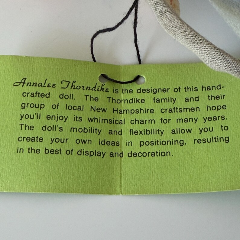 K&ouml;nnte beinhalten: Ein gr&uuml;nes Etikett mit schwarzem Text, auf dem steht: "Annalee Thorndike ist die Designerin dieser handgefertigten Puppe..." Das Etikett ist mit einer schwarzen Schnur an einer Puppe oder einem &auml;hnlichen Artikel befestigt. Der Text beschreibt die Eigenschaften der Puppe.