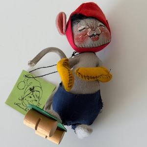 K&ouml;nnte beinhalten: Eine Vintage-Mauspuppe aus Filz mit roter Kapuze, blauer Hose und gelben Handschuhen. Die Puppe hat ein bemaltes Gesicht und h&auml;lt einen Holzschlitten. Ein gr&uuml;nes Etikett mit einer Zeichnung und dem Namen "Annalie" ist angebracht.
