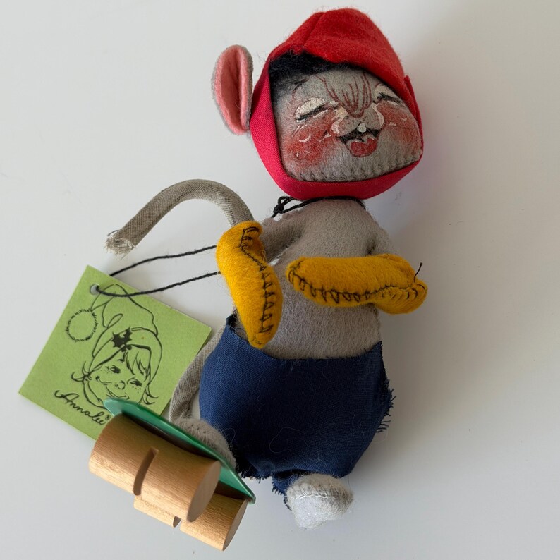K&ouml;nnte beinhalten: Eine Vintage-Mauspuppe aus Filz mit roter Kapuze, blauer Hose und gelben Handschuhen. Die Puppe hat ein bemaltes Gesicht und h&auml;lt einen Holzschlitten. Ein gr&uuml;nes Etikett mit einer Zeichnung und dem Namen "Annalie" ist angebracht.