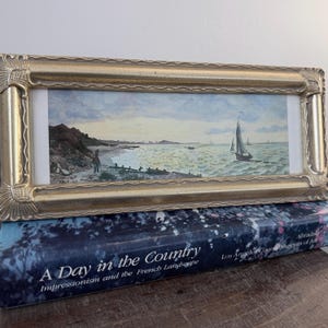 May include: A rectangular landscape painting in a decorative gold frame featuring a coastal scene with a sailboat on the water and a figure on the shore. The artwork is resting on a dark blue book titled A Day in the Country.