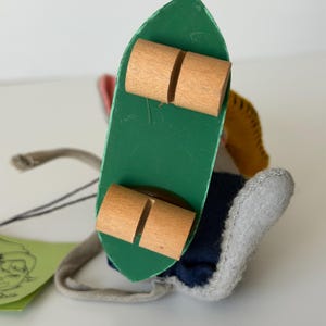 K&ouml;nnte beinhalten: Handgemachte Puppe mit einem gr&uuml;nen Skateboard mit Holzr&auml;dern. Die Puppe hat einen grauen Filzfu&szlig;, eine marineblaue Hose und ein gelbes und rotes Oberteil. Ein Anh&auml;nger mit einer Zeichnung ist befestigt.